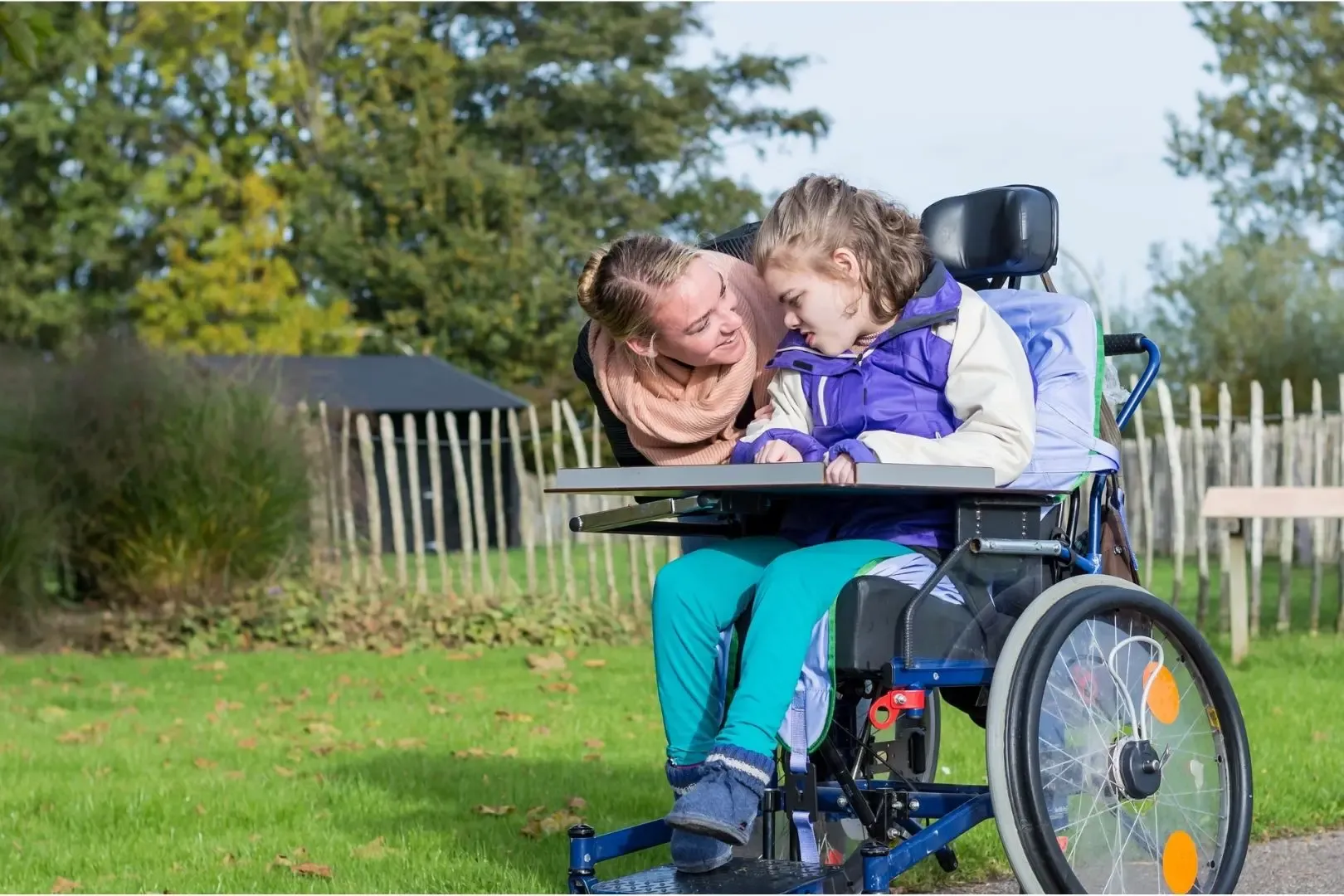 Assistentin beugt sich zu Kind im Rollstuhl und lächelt, draußen auf einer Wiese mit Bäumen.