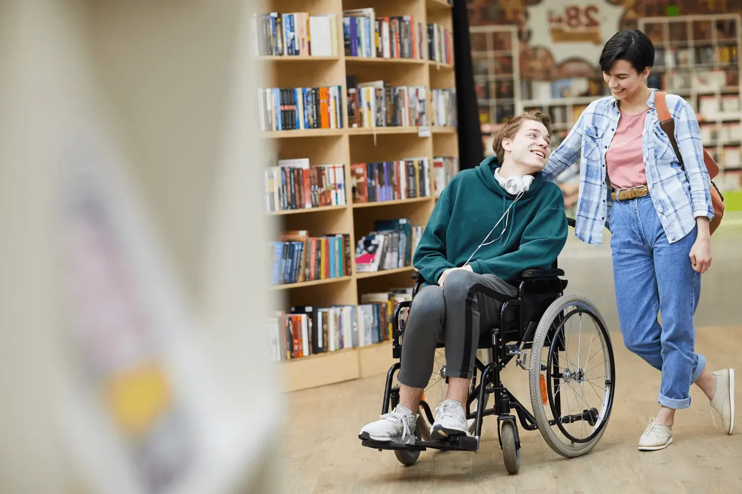 Junge Person im Rollstuhl wird von ihrer Assitentin in einer Bibliothek geschoben, Bücherregale im Hintergrund.