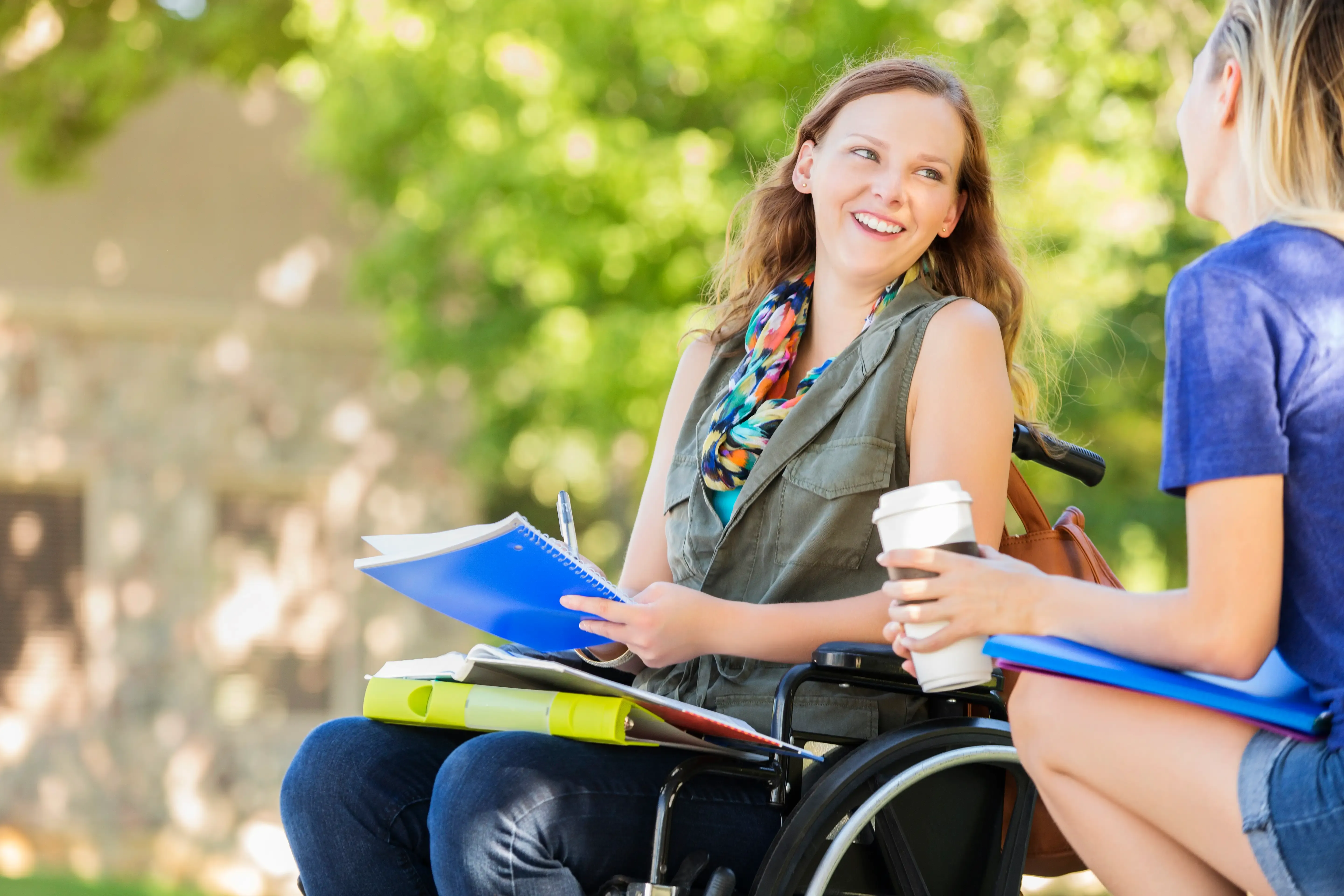 Frau im Rollstuhl mit Notizbüchern und Kaffeebecher spricht mit ihrer Assistentin im Park.