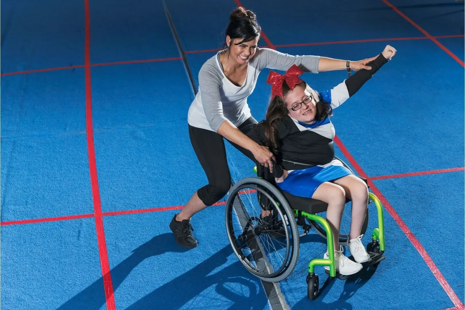 Sportassistentin unterstützt Mädchen im Rollstuhl beim Sport auf blauem Spielfeld mit roten Linien.