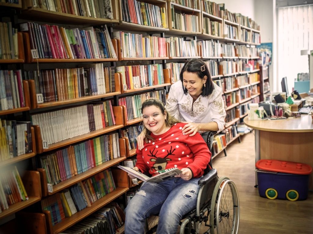 Ein Foto von einer Frau im Rollstuhl, die gemeinsam mit ihrer Assistentin in einer Bücherei ist.